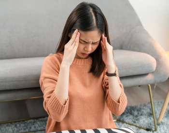 Female is sitting in front of a couch on the floor and making a facial expression of discomfort and holding her hands up to each side of her face